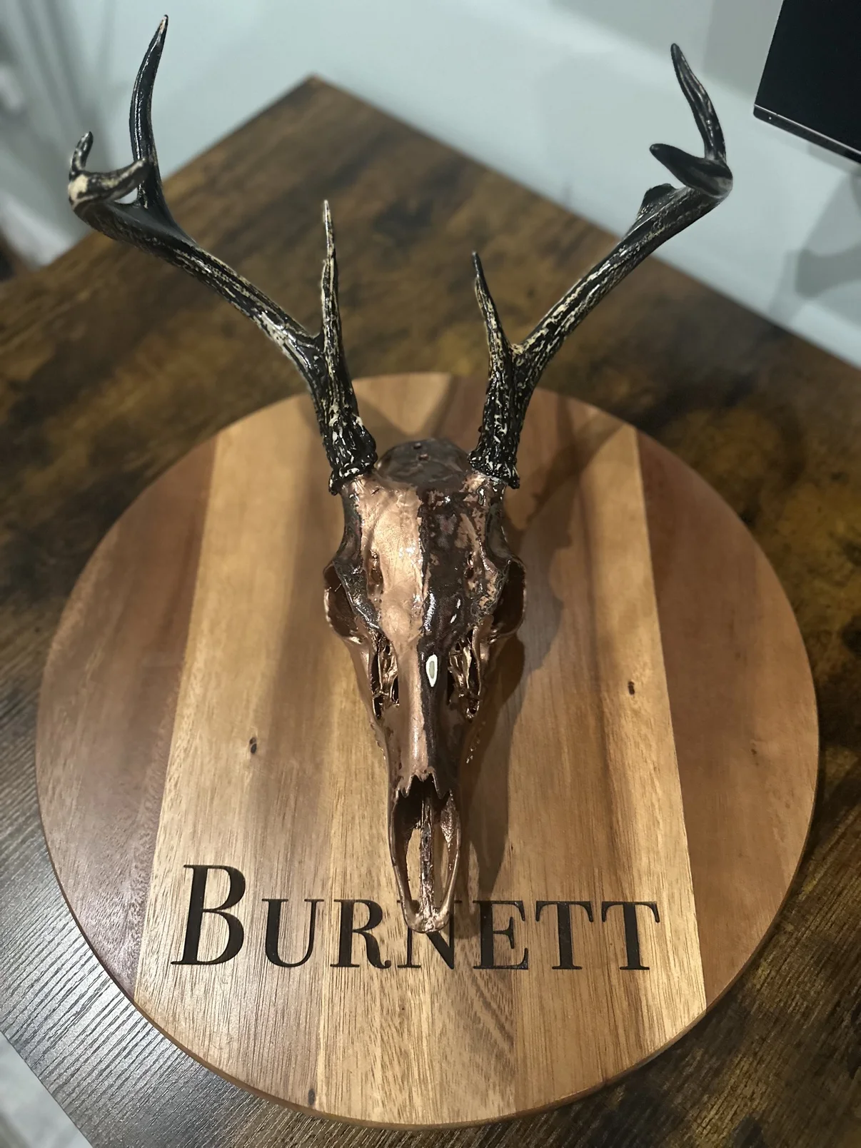 Photograph of my hand-painted deer skull from the front on a round wooden board, metallic copper finish with dark brown tones and black painted antlers