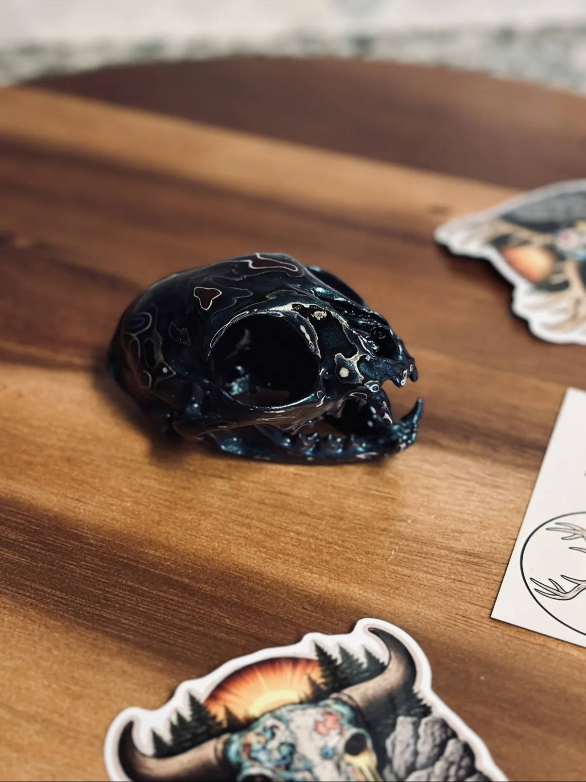 Photograph of my small hand-painted animal skull from an angled view on a wooden board, dark teal and black finish with red and white fordite accents, jaw attached
